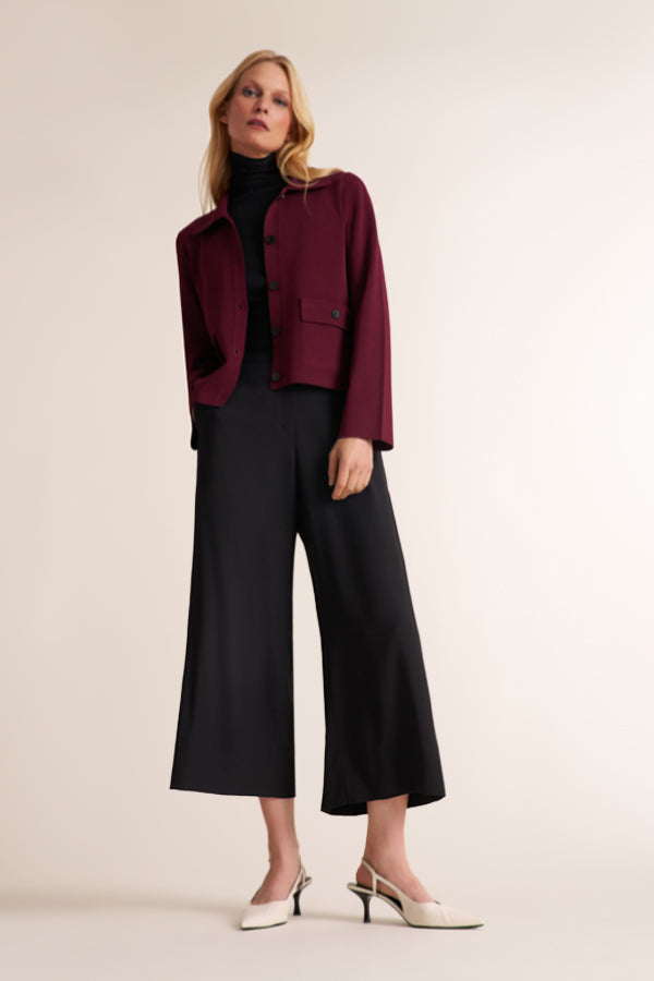 A woman modeling a cropped maroon jacket over a black turtleneck, paired with black wide-leg cropped pants and white pointed-toe heels, against a plain beige studio background. | Exclude